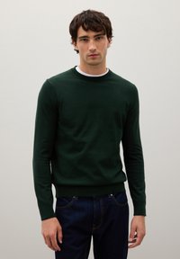 Dunkelgrüner Pullover mit langen Ärmeln und einem gerippten Ausschnitt. Glatte Textur, getragen über einem weißen Rundhalsausschnitt-Shirt.