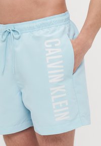 Lyseblå svømmeshorts laget av glatt stoff med elastisk midjebånd og hvit "CALVIN KLEIN" tekst på venstre side.