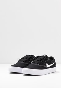 Une paire de baskets basses Nike noires avec des semelles blanches et des logos swoosh blancs, présentées sur une surface blanche contre un fond gris clair.