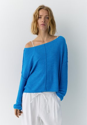 Jeune personne aux cheveux blonds mi-longs porte un pull bleu ample à épaules dénudées et un pantalon blanc à cordon, debout contre un mur blanc uni.