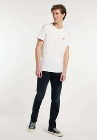 Camiseta de algodón blanca de manga corta, con un pequeño gráfico rojo en el lado izquierdo del pecho, combinada con vaqueros azul oscuro y zapatillas negras.