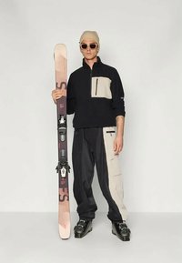 Completo da sci nero e beige con tasca con zip, che tiene uno sci con attacchi. Un berretto beige e occhiali da sole completano il look. Tessuto liscio.