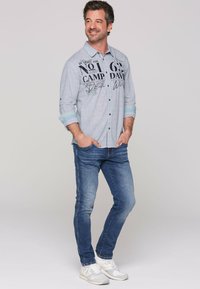 Camisa de botones azul claro con gráficos impresos, mangas remangadas, combinada con jeans ajustados azul oscuro y zapatillas blancas.