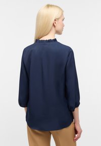 Blusa blu navy con scollatura a volant e maniche a tre quarti. Tessuto morbido e leggero. Abbinata a pantaloni beige.