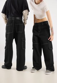 Black cargo pants with white stitching, multiple pockets, and a loose fit. Worn with contrasting footwear, showcasing a checkerboard pattern.
