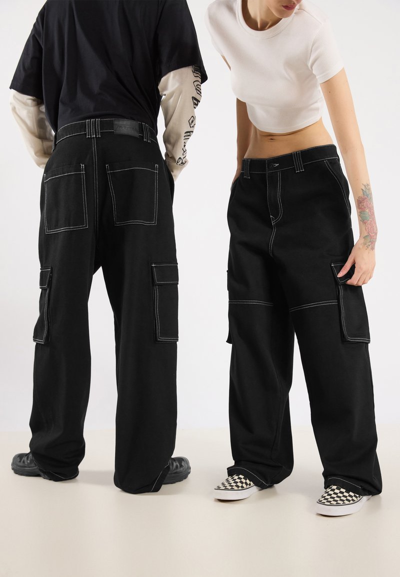 Black cargo pants with white stitching, multiple pockets, and a loose fit. Worn with contrasting footwear, showcasing a checkerboard pattern.