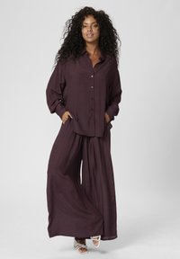 Dvojdielny outfit pozostávajúci z voľného, zapínacieho trička a nohavíc s širokými nohavicami, oboje v hlbokej farbe slivky s ľahkou textúrou.