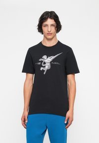 Mlad moški z valovitimi lasmi nosi črn Nike majico z angelom in besedilom, v kombinaciji s svetlo modrimi trenirkami, stoji pred belo ozadjem.
