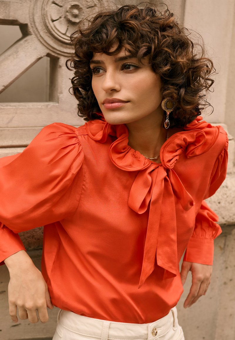 Blouse orange vif à manches bouffantes et col à volants, avec un détail nœud noué, assortie à un pantalon de couleur claire.