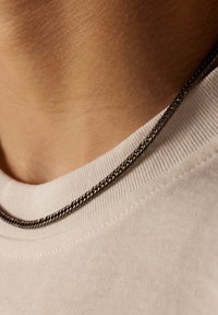 Zwarte kettinghanger rustend op een beige katoenen t-shirt, met de nadruk op een zachte textuur en een eenvoudig, verweven ontwerp.