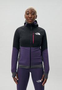 Fleece zip-jakke i svart og lilla med en tettsittende design, hette og logo på brystet. Har kontrasterende rosa glidelåsdetaljer.