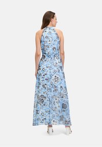 Maxi jurk in lichtblauwe chiffon met zwart en grijs bloemenpatroon, halternekontwerp en vloeiende, sheer stof. Dikke witte blokhakken.