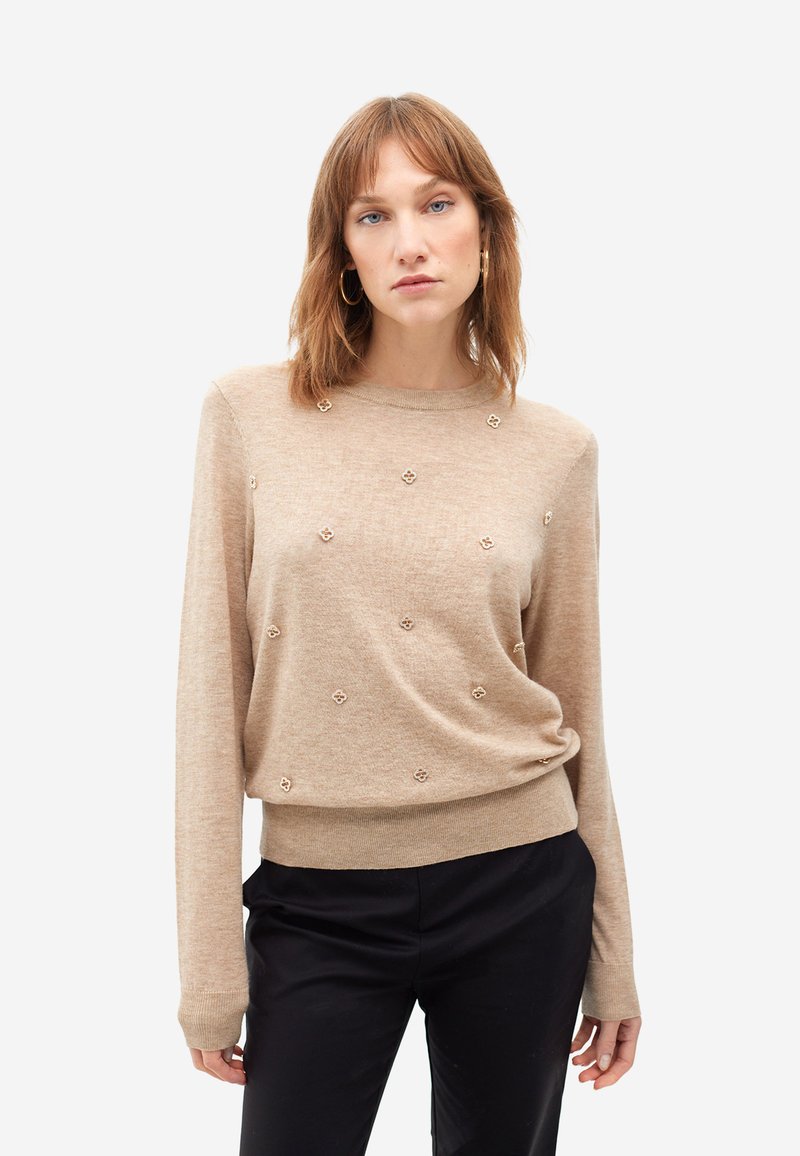 Jersey beige con mangas largas, escote redondo y apliques florales decorativos. Textura suave, corte relajado, combinado con pantalones negros.