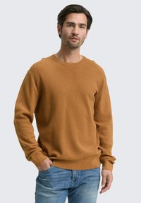 Suéter de punto marrón con cuello redondo y puños acanalados, que presenta un acabado texturizado. Llevado con jeans de color azul claro, manos en los bolsillos.