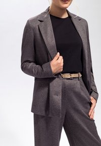 Grauer maßgeschneiderter Blazer mit subtiler Textur, kombiniert mit passenden Hosen. Getragen über einem schwarzen Oberteil und akzentuiert durch einen hellen Gürtel.
