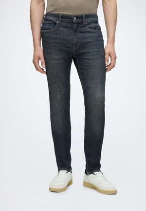 Man draagt slim fit donkerblauwe jeans en witte sneakers, staand tegen een effen achtergrond.