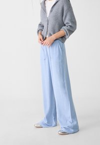 Pantalons larges bleu clair avec une taille à cordon, fabriqués dans un tissu doux et fluide. Associés à un cardigan texturé gris et des chaussures argentées.