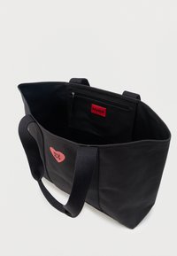 Μαύρη τσάντα tote με δύο λαβές, κόκκινο λογότυπο καρδιάς με τη λέξη "HUGO" στο μπροστινό μέρος και εσωτερική τσέπη με φερμουάρ και κόκκινη ετικέτα "HUGO".