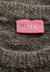 Tissu tricoté brun, épais et doux, avec une étiquette de vêtement rose cousue, affichant le nom de la marque JJXX en lettres blanches.