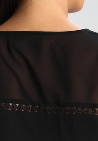 Zwarte top met een doorzichtige rugpaneel en een cirkelvormige kanten randafwerking. Textuurstof creëert een gelaagd uiterlijk. Eenvoudig ontwerp van de halslijn.