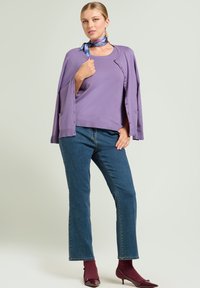 Top morado de punto bajo un cárdigan abierto a juego, combinado con vaqueros azules y tacones de charol burdeos. La bufanda presenta patrones azules y rosas.