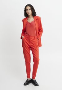 Red suit with fitted trousers and blazer, layered over a red top with lace detailing. Black loafers complete the outfit, offering a sleek look.