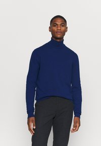 Marinefarbener Rollkragenpullover aus weichem Material, mit langen Ärmeln und taillierter Passform. Kombiniert mit dunkel strukturierten Hosen.