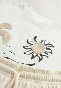Maglietta bianca in cotone con stampa floreale nera, completata da un accento testurizzato a forma di sole beige e un bordo in maglia color crema nella parte inferiore.