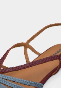Σανδάλια με πλεκτές λουρίδες σε μπλε, μαρόν και ταμπά χρώματα σε ουδέτερο φόντο, με κοντινή λήψη της επιφάνειας του ποδιού και των λουριών.