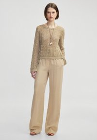 Beige, gestrickter Langarm-Top mit einem strukturierten Wellenmuster, kombiniert mit locker sitzenden, hochgeschnittenen beigen Hosen; minimalistische Accessoires.