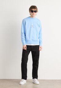 Felpa azzurra con scollo rotondo, caratterizzata da un piccolo logo ricamato. Abbinata a pantaloni neri e sneakers bianche. Indossati occhiali da sole neri.
