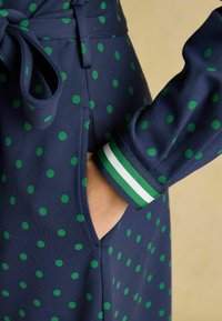 Jūras zila kleita ar lieliem zaļiem punktiņiem, ar auklu jostu, sānu kabatām un svītrainiem zaļiem un baltiem akcentiem uz piedurknēm.