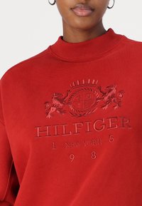 Crvena dukserica s visokim rebrastim ovratnikom. Sadrži urezani logo s lavovima i natpis "HILFIGER 1 NEW YORK 1985". Glatka tekstura.