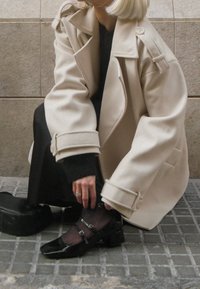 Personne aux cheveux blond clair portant un manteau beige et une robe noire ajustant des chaussures Mary Jane noires à talons sur un trottoir carrelé gris.