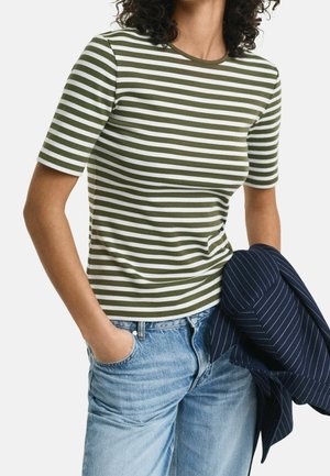Mujer vestida con una camiseta de manga corta a rayas verdes y blancas con vaqueros azul claro, sosteniendo una chaqueta azul marino de rayas finas en una mano.