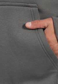 Grauer Hoodie-Stoff mit weicher Textur, der eine Fronttasche aufweist, in die die Hand teilweise eingefügt ist. Die Stickdetails sind entlang der Kanten sichtbar.