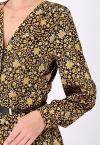 Robe noire à fleurs avec des motifs jaunes et blancs, manches longues, décolleté en V profond et taille cintrée avec un détail de ceinture noire.