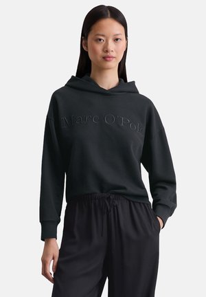 Schwarzer Hoodie aus weichem Material mit einem Logo-Text auf der Vorderseite. Verfügt über eine lockere Passform, seitliche Taschen und eine Kapuze mit Kordelzug. Kombiniert mit schwarzen Hosen.