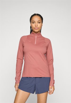 Woman with braided hair wearing a long-sleeve, rose-colored zip-up top and navy athletic shorts, standing against a plain light background.