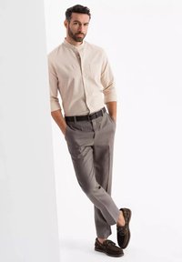 Jasno beżowa koszula zapinana na guziki, szare dopasowane spodnie, czarny pasek, brązowe buty typu boat shoes. Model opiera się o jasne tło.