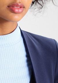 Detailní záběr na krk a bradu osoby, na které je vidět světle modrý žebrovaný trikot s vysokým nákrčníkem pod tmavě modrým sakem. Hladká pleť a výrazné rty.