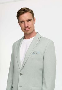 Hellgrauer Blazer aus strukturiertem Stoff, mit Revers, einem Knopfverschluss und einem Einstecktuch mit blau-weißen Mustern. getragen über einem weißen Hemd.