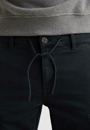 Nahaufnahme von schwarzen Hosen mit einem Knopf, einem zu einer Schleife gebundenen Kordelzug und einem grauen, gerippten Pullover, der leicht in den Hosenbund gesteckt ist.