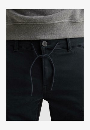 Detailní záběr černých kalhot s knoflíkem, šňůrkou zavázanou na mašli a šedého žebrovaného svetru mírně zastrčeného do pasu.