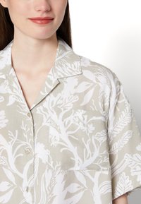 Camisa de manga curta em bege claro com padrão botânico branco. Apresenta um colarinho e um bolso no peito. O tecido parece suave e leve.