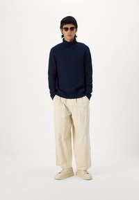 Suéter de punto acanalado azul marino, pantalones sueltos color crema y zapatillas crema. El conjunto presenta formas relajadas con acentos mínimos.
