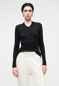 Camisola preta de malha canelada com decote em V e mangas compridas, apresentando uma silhueta ajustada e fendas laterais, combinada com calças de cor clara.