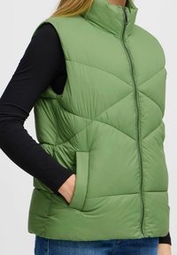 Zeleni puffer prsluk s visokim ovratnikom, zatvorenjem na zip, dijamantnim kvadratnim uzorkom i bočnim džepovima, složen preko crnog dukserice s dugim rukavima.