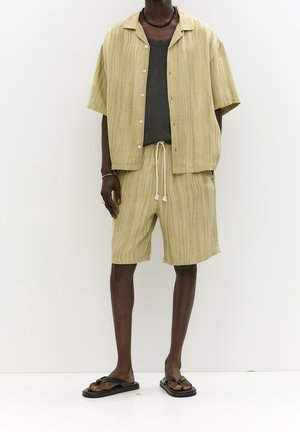 Camisa de manga corta color beige claro y pantalones cortos con rayas verticales, combinados con una camiseta sin mangas oscura y chanclas negras. Material de algodón.
