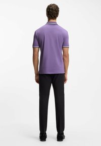 Polo viola con colletto a righe bianche e gialle; abbinata a pantaloni e scarpe nere, vista da dietro. Maniche corte, design aderente.